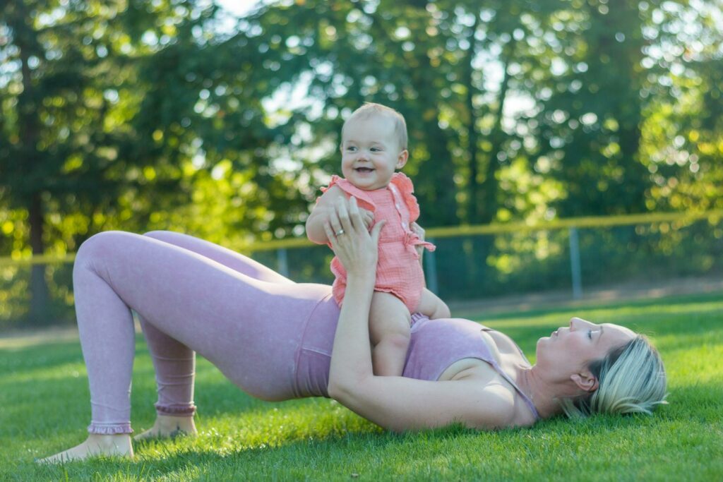 A mother in activewear exercises with her baby in a sunny park, promoting a healthy lifestyle.