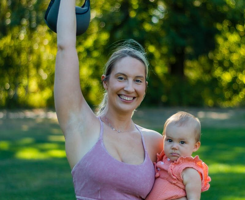 Smiling woman exercising outdoors with baby, holding a kettlebell.